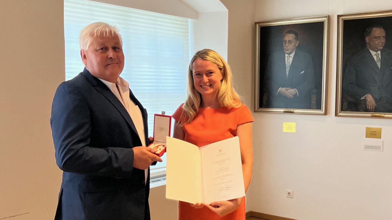 Ein Mann und eine Frau stehen in einem Raum, der Mann hält eine Medaille und die Frau ein Zertifikat. Sie lächeln und scheinen eine Leistung zu feiern. Hinter ihnen hängt ein Porträt eines Mannes in Anzug.