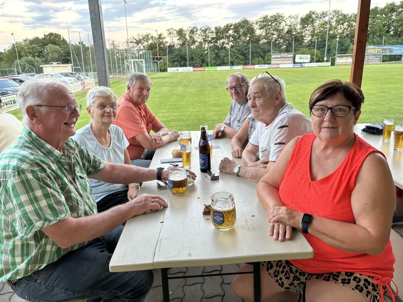 A group of elderly people sitting around a table at a sports stadium, enjoying drinks and watching a game.