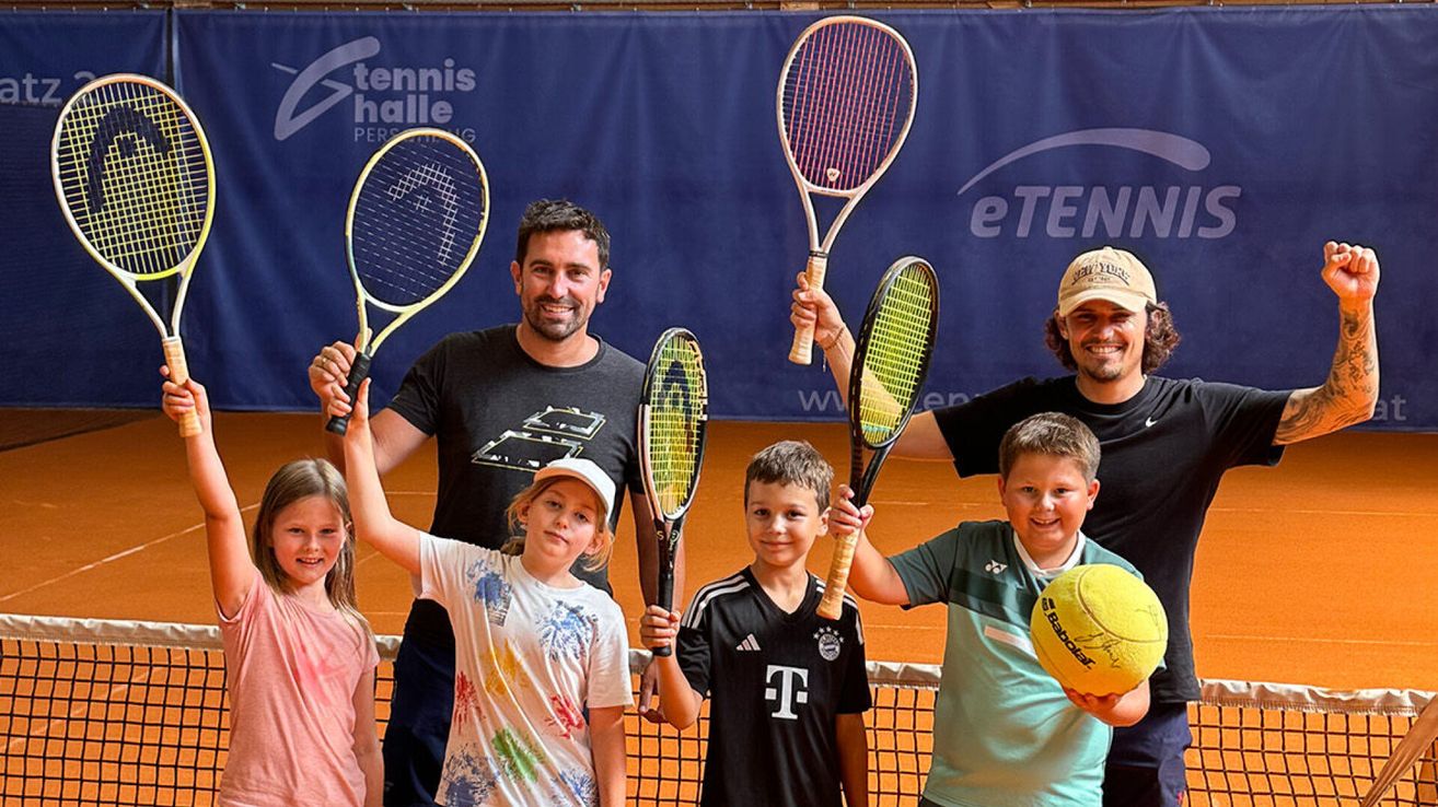 Four individuals, two adults and two children, are standing on a tennis court holding tennis rackets. The adults are smiling and appear to be posing for a photo. The children are holding tennis balls.