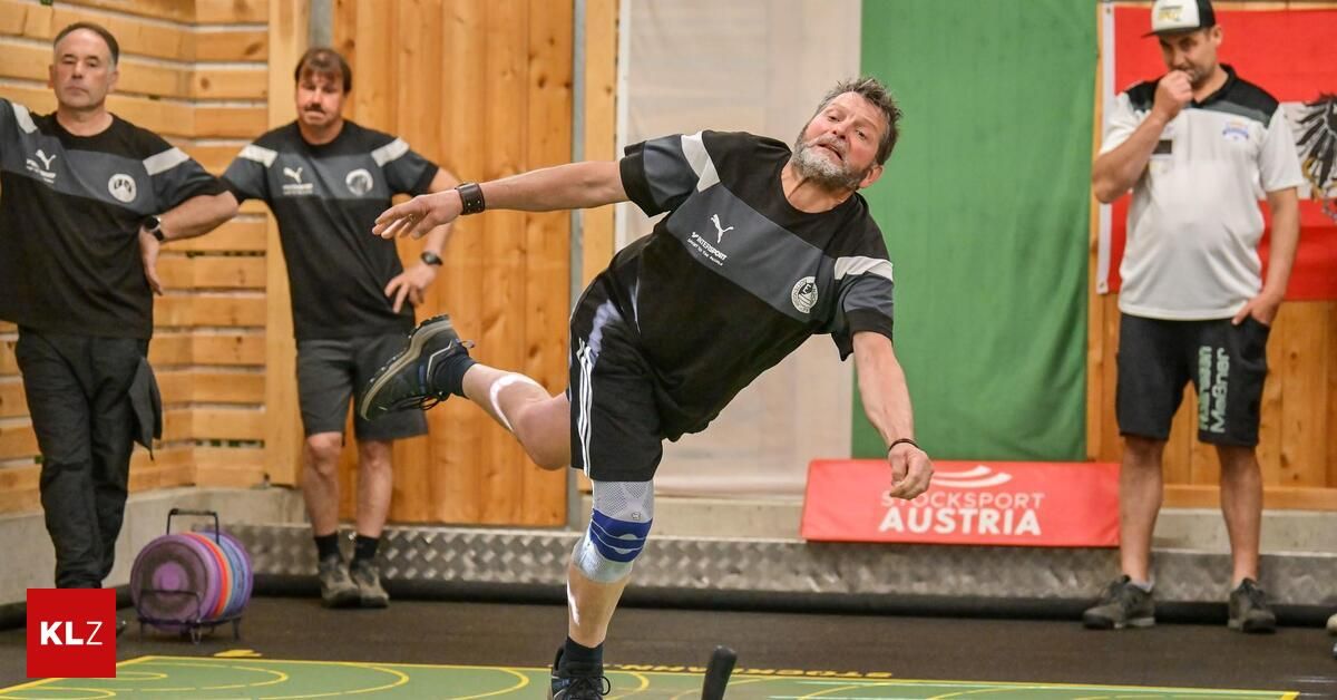 A man in a black and gray sports uniform kicks a ball in an indoor sports facility. Another man stands behind him, watching. The wall behind them is wooden, and there is a green banner with white text.
