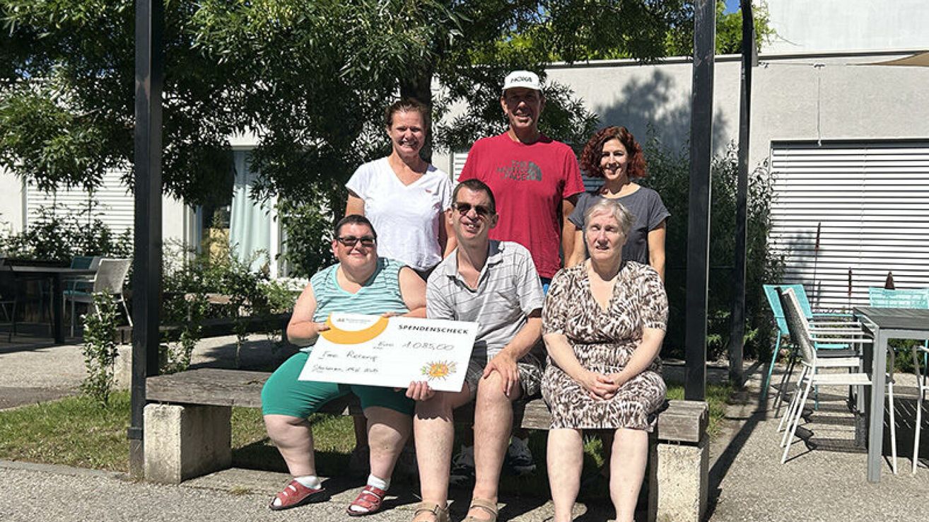 A group of six people are sitting on a bench, smiling and posing for a photo. One person is holding a check and a sign.