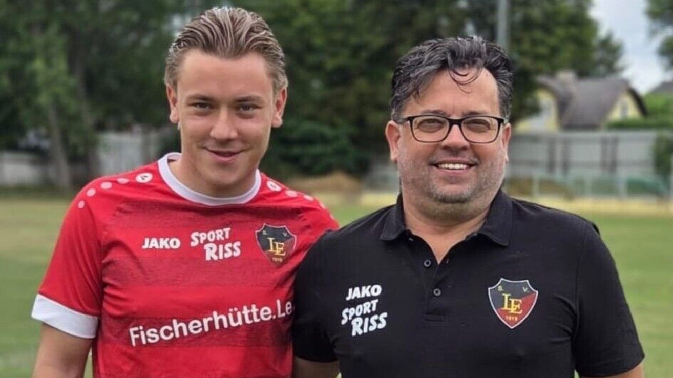 Two men standing side by side, one in a red sports jersey with 'SPORT RISS' and the other in a black polo with 'Jako Sport RISS'. They are smiling and appear to be posing for a photo.