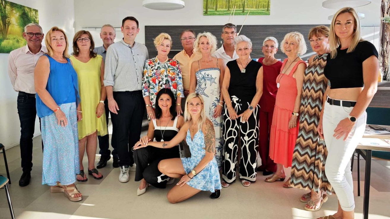Eine Gruppe von Menschen, darunter Männer und Frauen, posiert für ein Foto in einem Raum mit weißer Wand und Bilderrahmen.