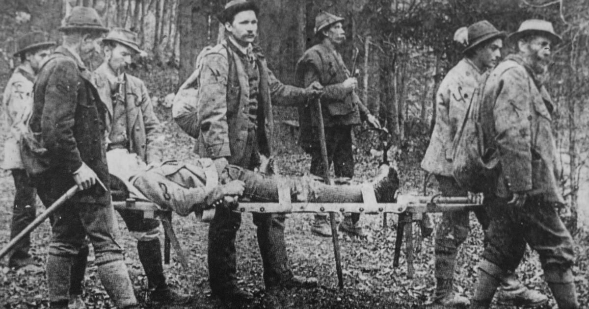 A black and white image of soldiers carrying an injured comrade on a stretcher through a forest.