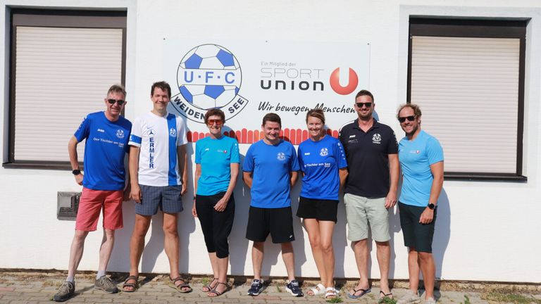 A group of people in matching blue shirts and shorts are standing in front of a white wall with a logo and text.