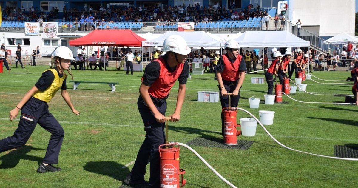 Eine Gruppe von Personen in roten Westen und weißen Helmen übt Feuersicherheitsübungen auf einem Feld mit Zuschauern im Hintergrund.