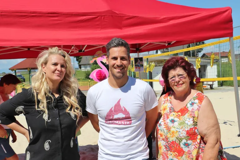 Drei Personen stehen unter einem roten Zelt und lächeln für ein Foto. Der Mann trägt ein weißes T-Shirt mit Logo, während die Frauen schwarze und blumige Kleider tragen. Dahinter sind eine pinke Flamingo-Dekoration und ein gelbes Netz zu sehen.