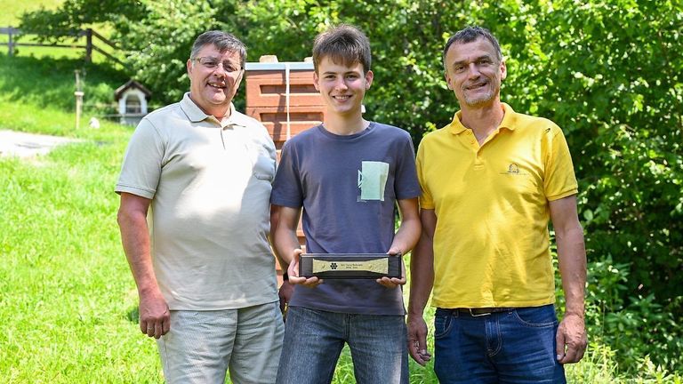 Drei Männer stehen in einem Feld und lächeln. Der Mann in der Mitte hält eine Trophäe. Hinter ihnen steht eine Ziegelstruktur.