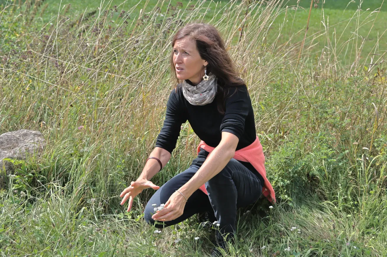Eine Frau kniet in einem Grasfeld, hält eine kleine weiße Blume und scheint etwas zu erklären.