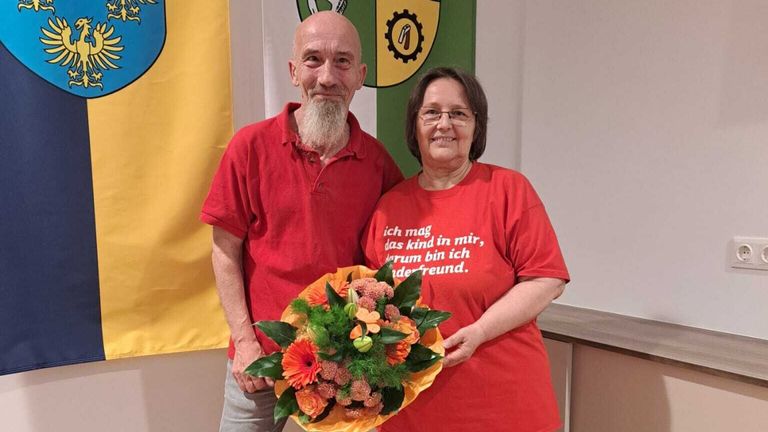 Ein älterer Mann und eine Frau stehen lächelnd nebeneinander, mit einem Blumenstrauß vor ihnen. Der Mann hat einen weißen Bart und die Frau trägt eine Brille. Hinter ihnen ist eine grün-gelbe Fahne.