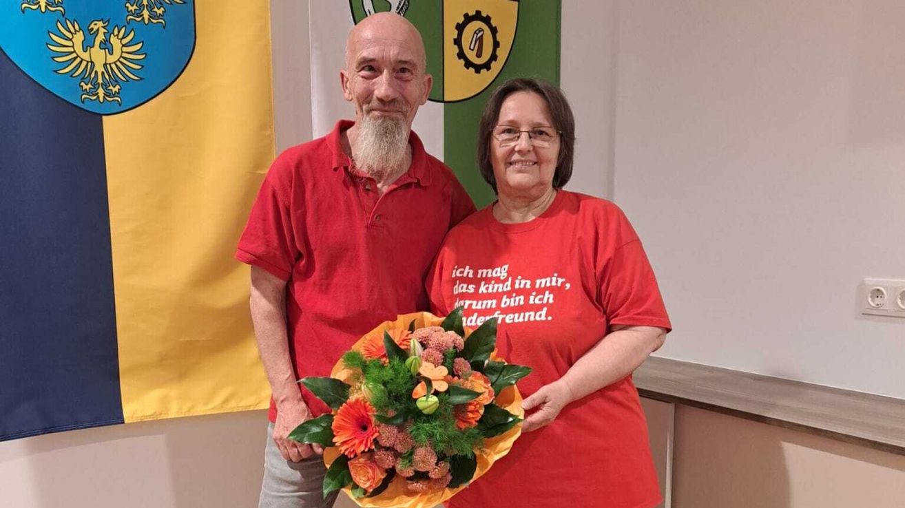 Ein älterer Mann und eine Frau stehen lächelnd nebeneinander, mit einem Blumenstrauß vor ihnen. Der Mann hat einen weißen Bart und die Frau trägt eine Brille. Hinter ihnen ist eine grün-gelbe Fahne.