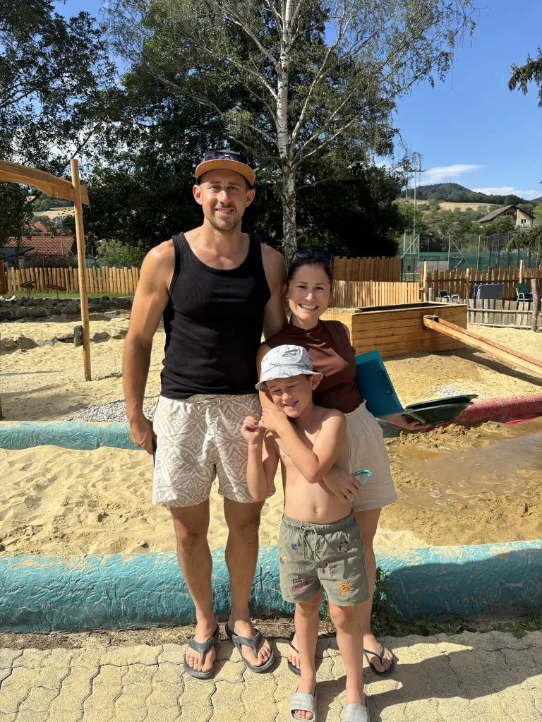 A man, a woman, and a young boy stand together in a sandy area, possibly at a playground. The man wears a black tank top and shorts, while the woman holds a shovel. The boy wears a hat and has his hand on his chin.