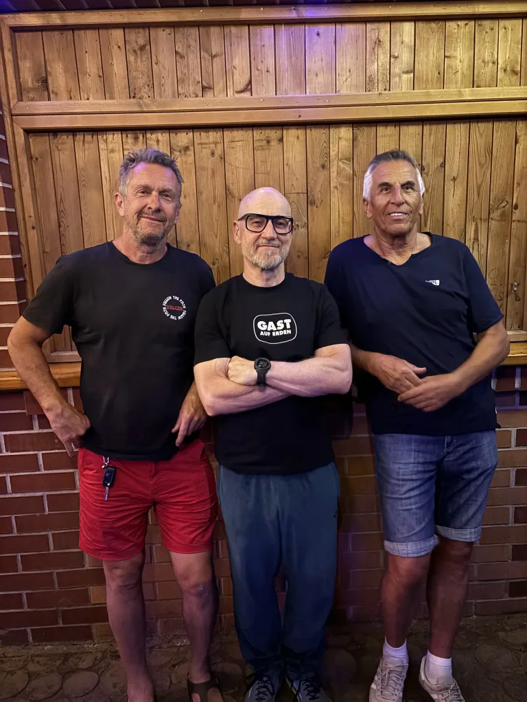 Drei Männer stehen gemeinsam vor einer Holzwand, lächeln und posieren für ein Foto. Der Mann in der Mitte trägt eine Brille und eine Uhr, während der Mann rechts einen Schlüsselanhänger hat.