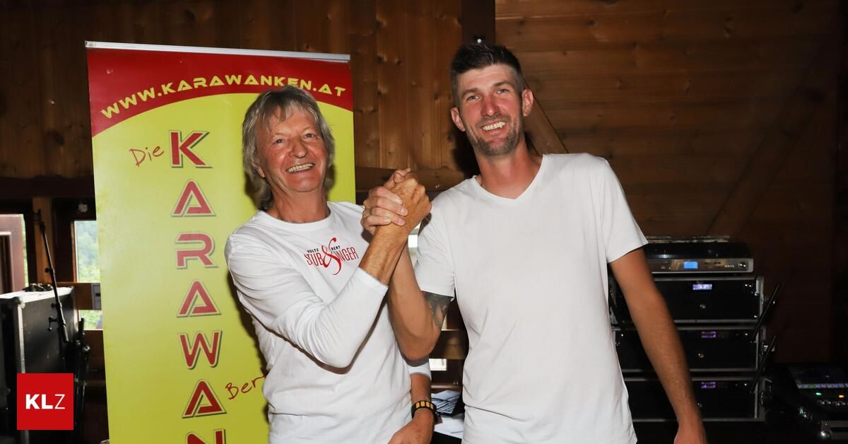 Two men standing and smiling in front of a wooden wall with a banner that reads 'Karawanken.at'. The man on the left wears a white shirt with red text and the man on the right has a tattoo on his arm.