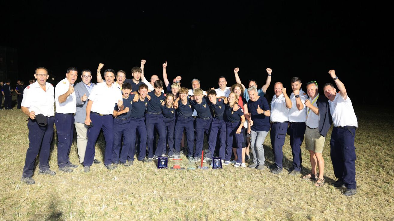 A group of people in navy blue uniforms and white shirts are posing for a photo on a grassy field at night. They are holding trophies and raising their hands.