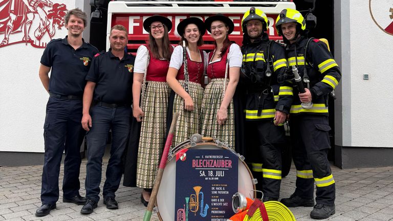 A group of people in traditional Bavarian attire, including three women with musical instruments, stand beside a fire truck. Two men in firefighter uniforms stand on either side of the group. The women are smiling, and the men are looking at the camera. A drum and a poster are in front of them.