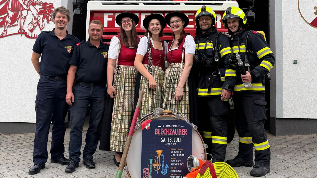 A group of people in traditional Bavarian attire, including three women with musical instruments, stand beside a fire truck. Two men in firefighter uniforms stand on either side of the group. The women are smiling, and the men are looking at the camera. A drum and a poster are in front of them.