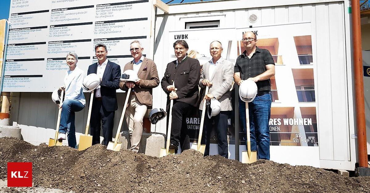 A group of men and women stand in front of a building with shovels in hand, wearing hard hats and smiling for a photo. Behind them is a wall with some text and images.