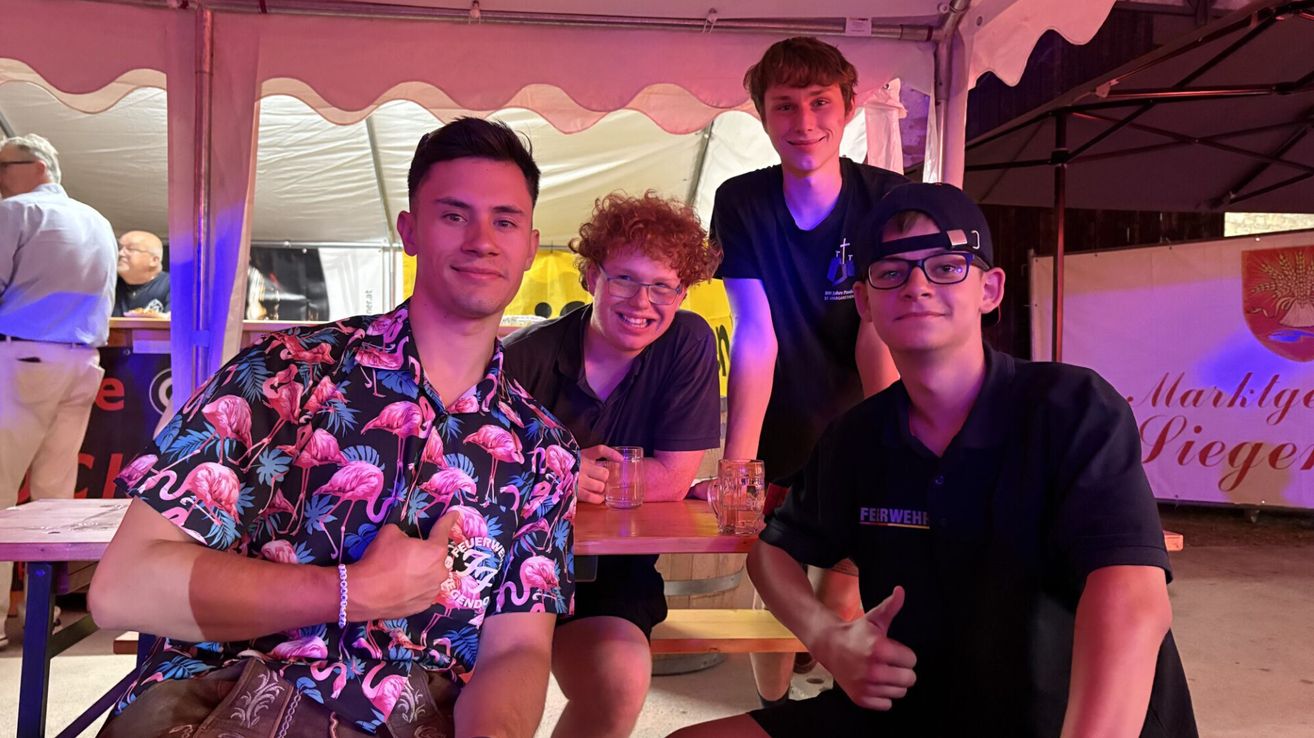 Four young men are smiling and posing for a photo in a tent with a pink light. The man on the left is wearing a floral shirt. The man in the middle is wearing glasses and a black shirt. The man on the right is wearing a black shirt with the word 'Feuerwehr' on it.