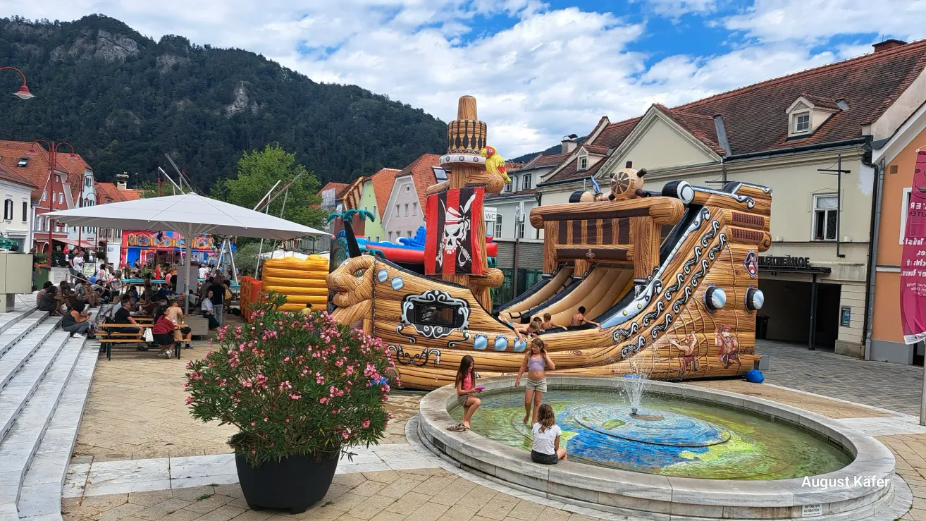 Kinder spielen in einem Brunnen neben einem großen aufblasbaren Piratenschiff auf einem Stadtplatz, mit Bergen im Hintergrund.