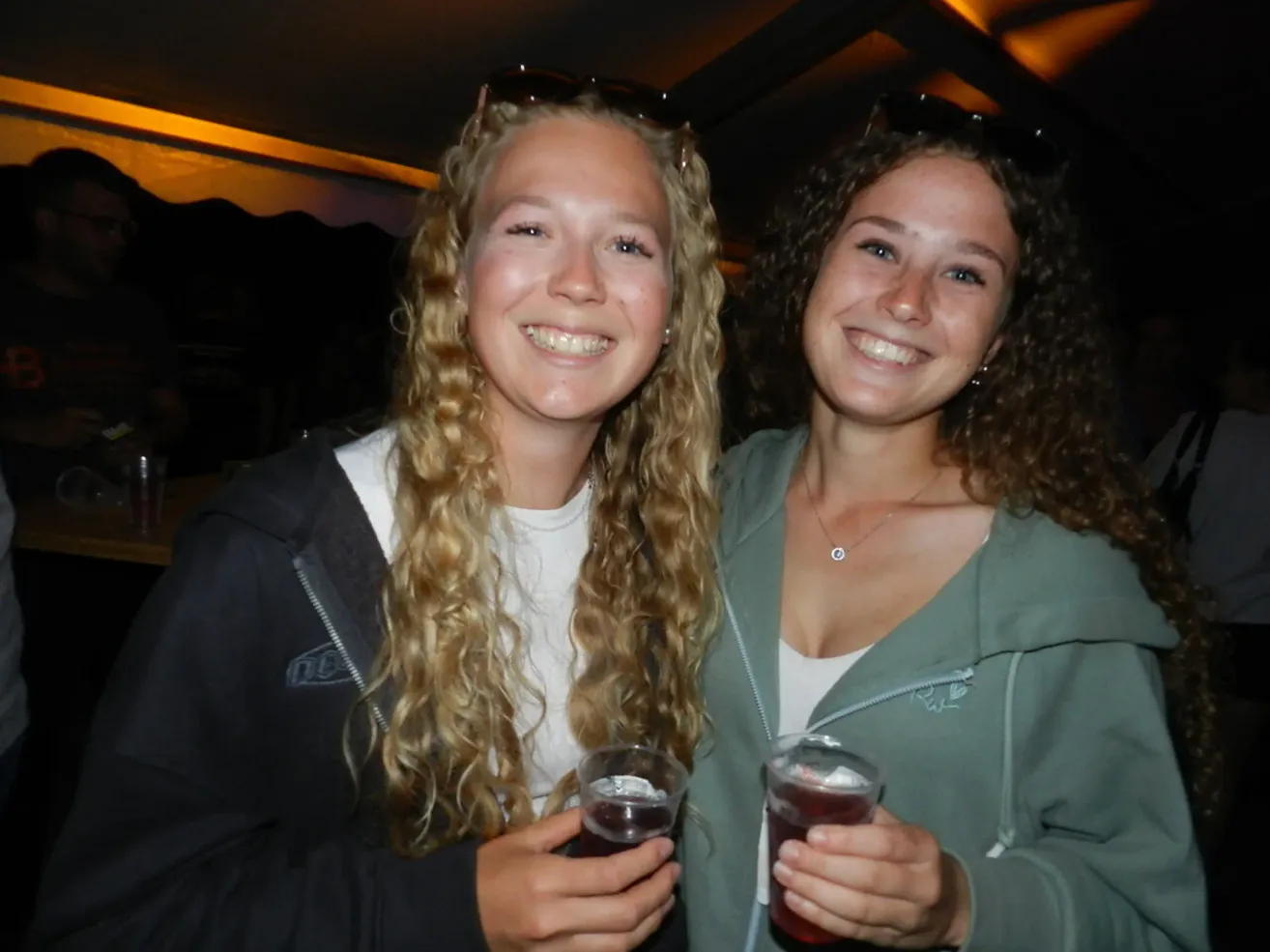 Zwei Frauen mit langen lockigen Haaren lächeln und posieren für ein Foto, während sie Getränke in den Händen halten.