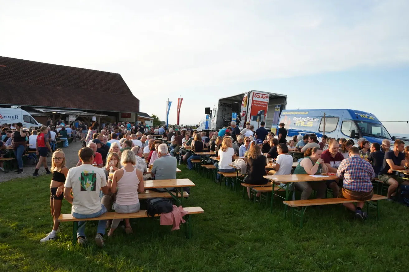 A large crowd of people are gathered in an outdoor area, sitting on benches around tables, possibly at a festival. A building with a flag is in the background.