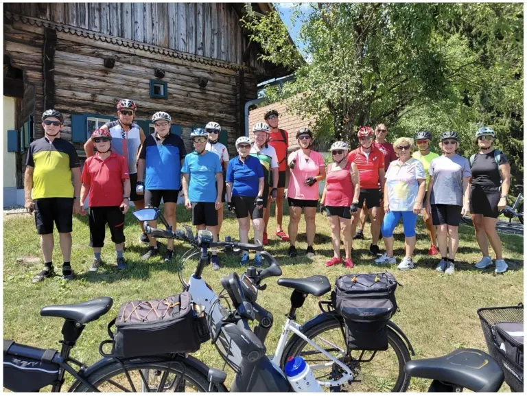 Eine Gruppe von Radfahrern mit Helmen und Fahrradausrüstung steht vor einem Holzhaus mit geparkten Fahrrädern.