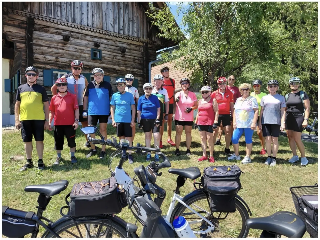 Eine Gruppe von Radfahrern mit Helmen und Fahrradausrüstung steht vor einem Holzhaus mit geparkten Fahrrädern.