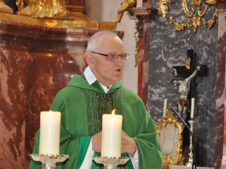 Ein älterer Priester in einer grünen Robe steht vor Kerzen, möglicherweise während einer religiösen Zeremonie. Hinter ihm befindet sich eine Wand mit einem goldenen Kreuz und einer Statue.