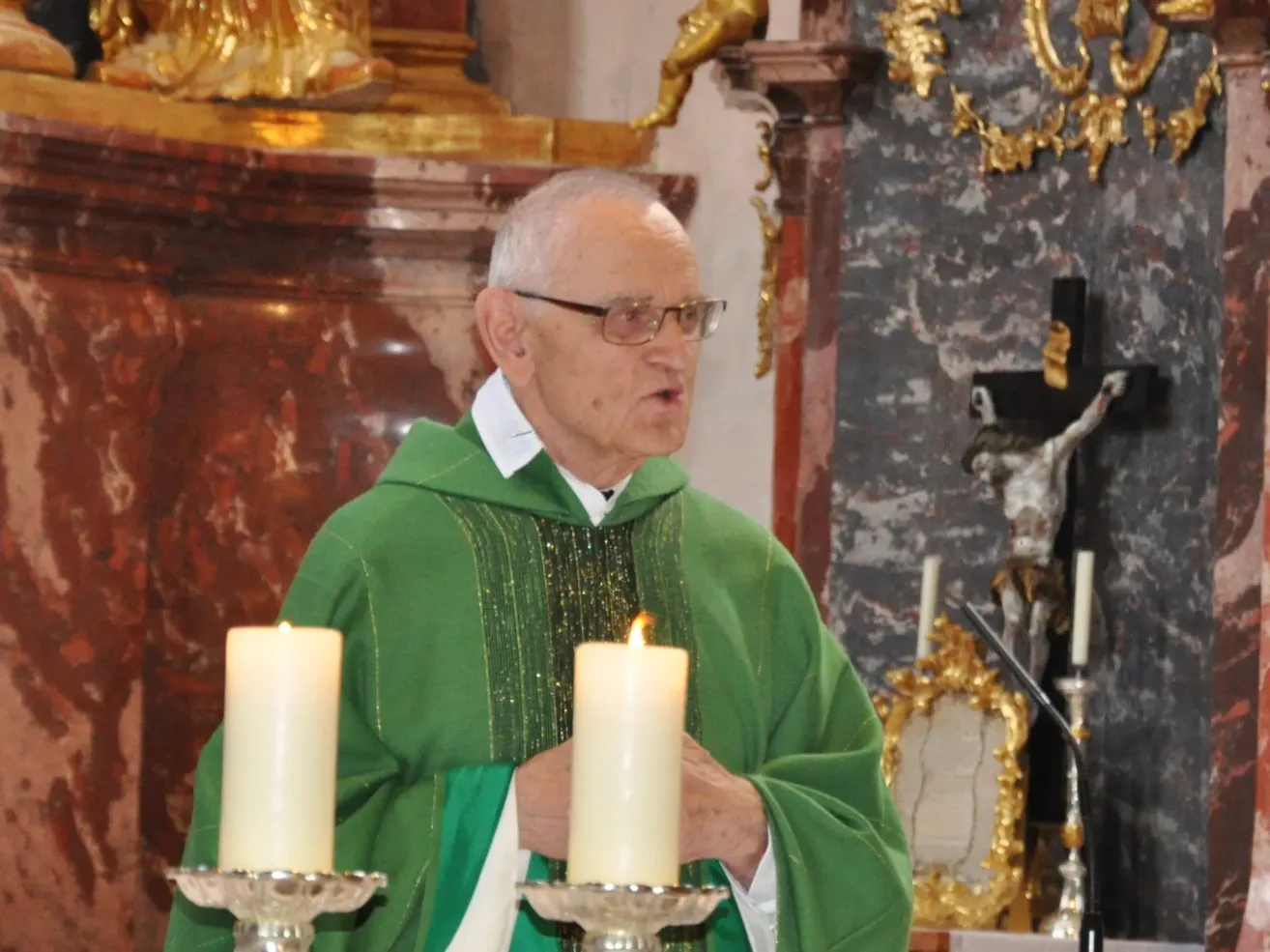 Ein älterer Priester in einer grünen Robe steht vor Kerzen, möglicherweise während einer religiösen Zeremonie. Hinter ihm befindet sich eine Wand mit einem goldenen Kreuz und einer Statue.