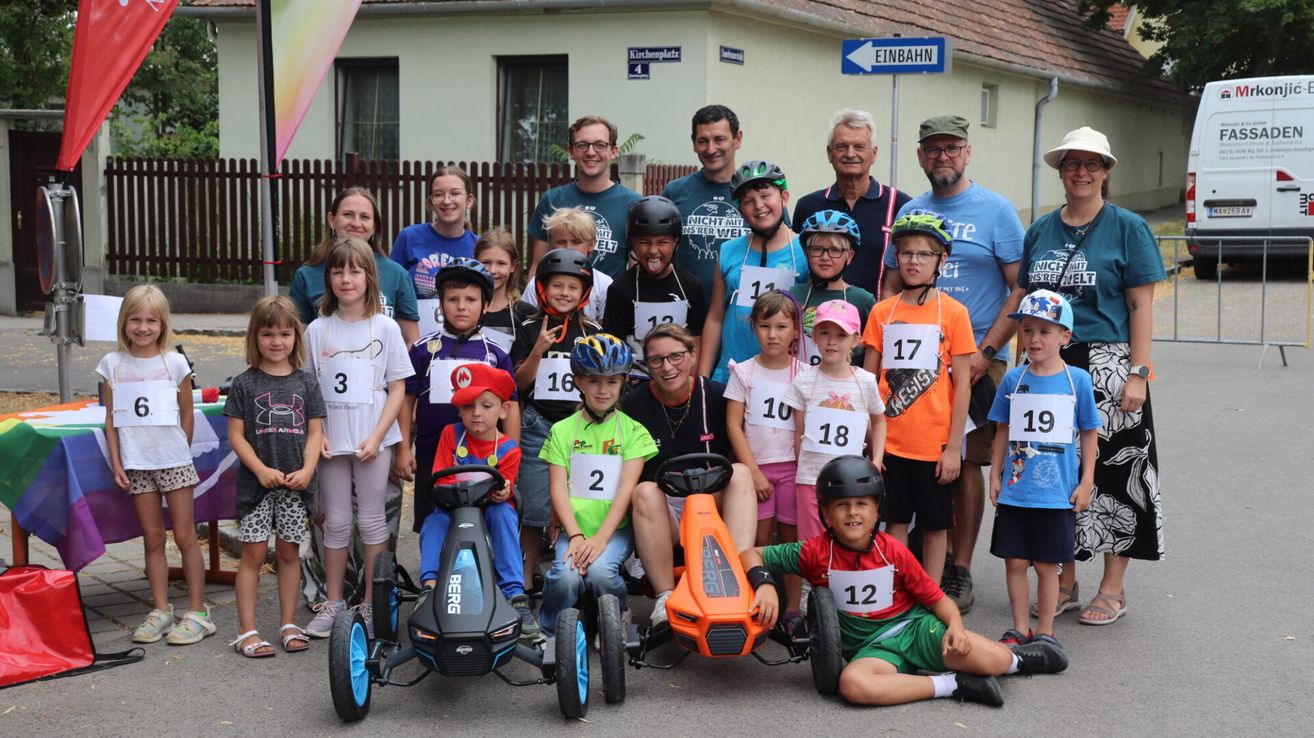 A group of children and adults are posing for a photo in front of a building with a sign that says Einbahn. The children are wearing helmets and some are sitting on go-karts. They are all wearing numbers on their shirts.