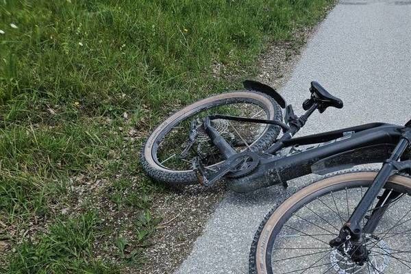 Ein schwarzes Fahrrad liegt auf der Seite eines gepflasterten Weges, mit einem losen Rad und einem verbogenen Rahmen. Das Fahrrad ruht auf Gras und Kies.