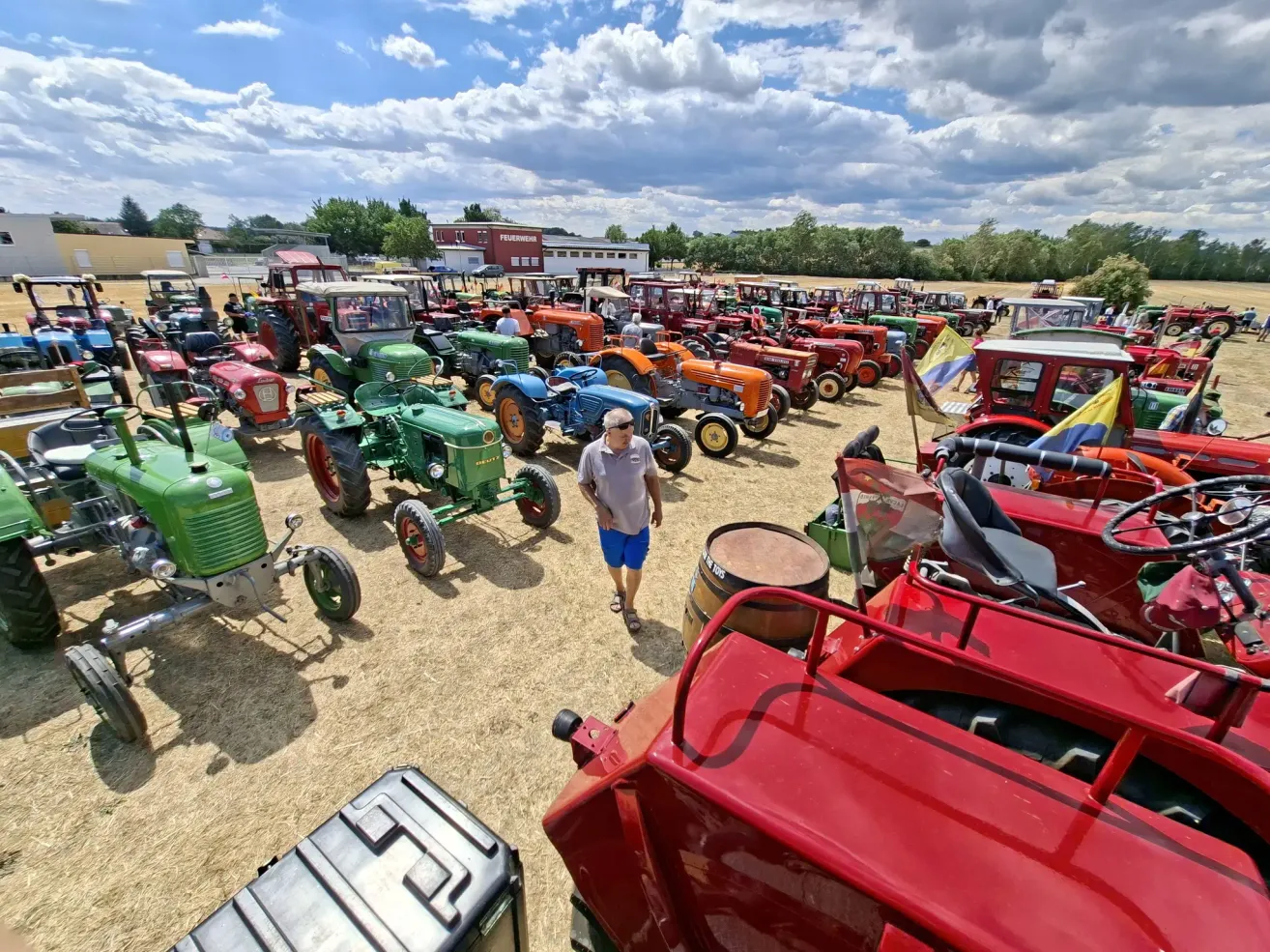A man walks past a large collection of vintage tractors parked in a field under a partly cloudy sky.