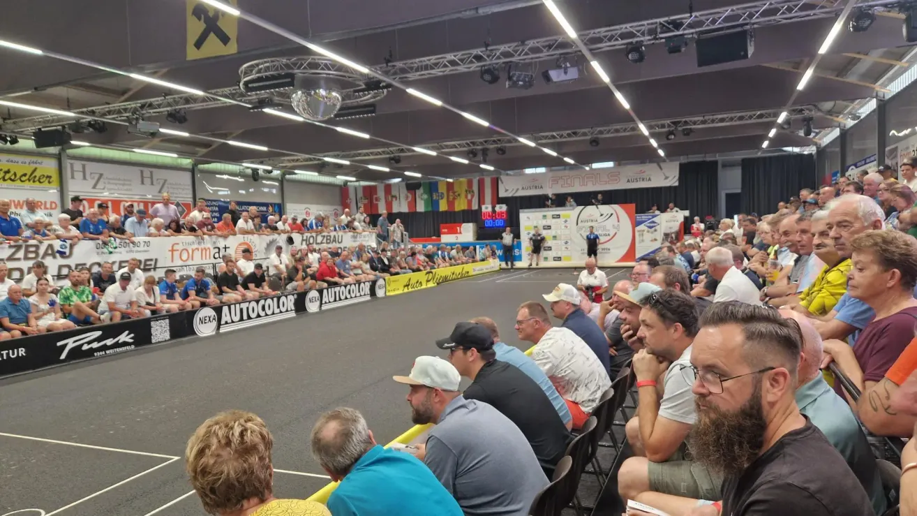 A crowd of spectators watches a sports event in an indoor arena. The audience sits in rows, and the court is visible with banners and flags. The scoreboard is displayed prominently.