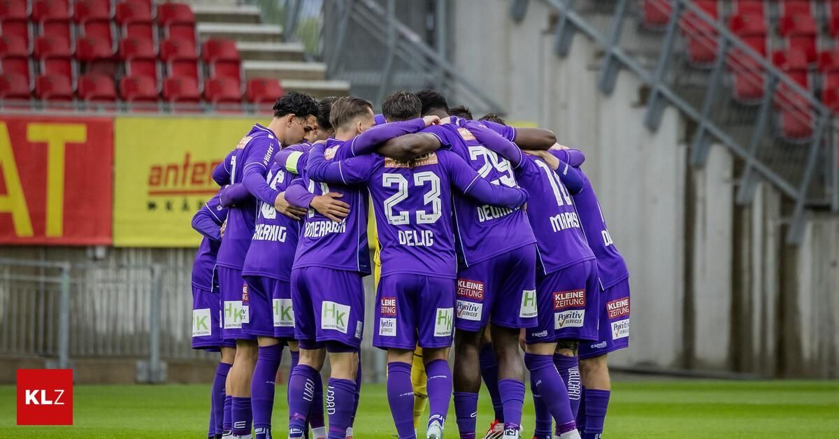 A soccer team in purple uniforms is huddled together on a field, possibly preparing for a game. The players are standing in a circle with their arms around each other.