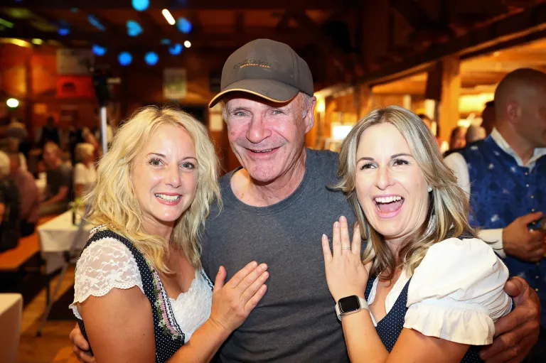 A man wearing a baseball cap and a gray t-shirt is smiling and posing for a photo with two women, one of whom is wearing a polka-dot dress and a watch.