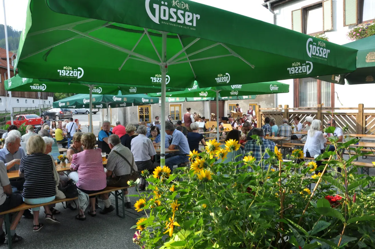 Eine Gruppe von Menschen sitzt unter grünen Schirmen an Tischen in einem Restaurant im Freien. Einige trinken Bier und eine Band spielt im Hintergrund.