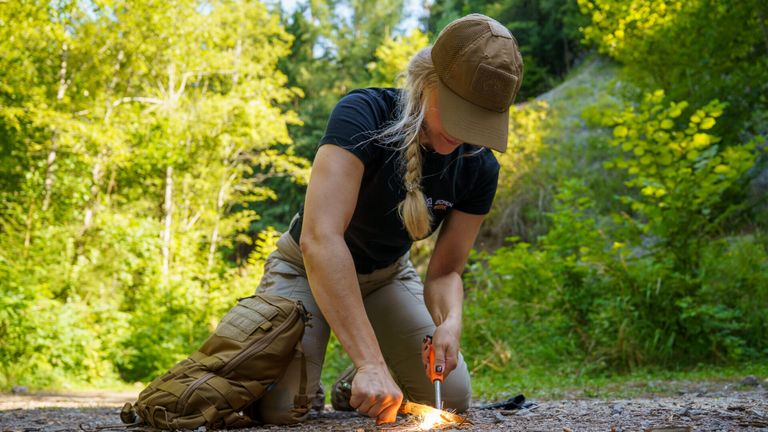 Eine Frau in einem schwarzen Shirt und khakifarbener Hose kniet auf dem Boden und benutzt ein Werkzeug, um ein Feuer zu entfachen. Sie trägt einen braunen Hut und hat einen Rucksack in der Nähe.