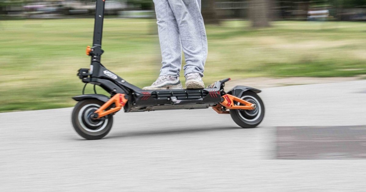 A person is riding a black and orange electric scooter on a paved road with a blurred background.