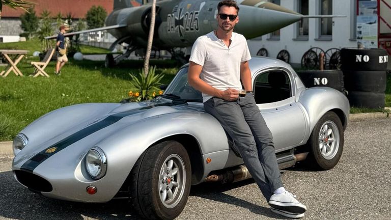 A man sits on a silver sports car, holding a drink, with a vintage jet in the background.