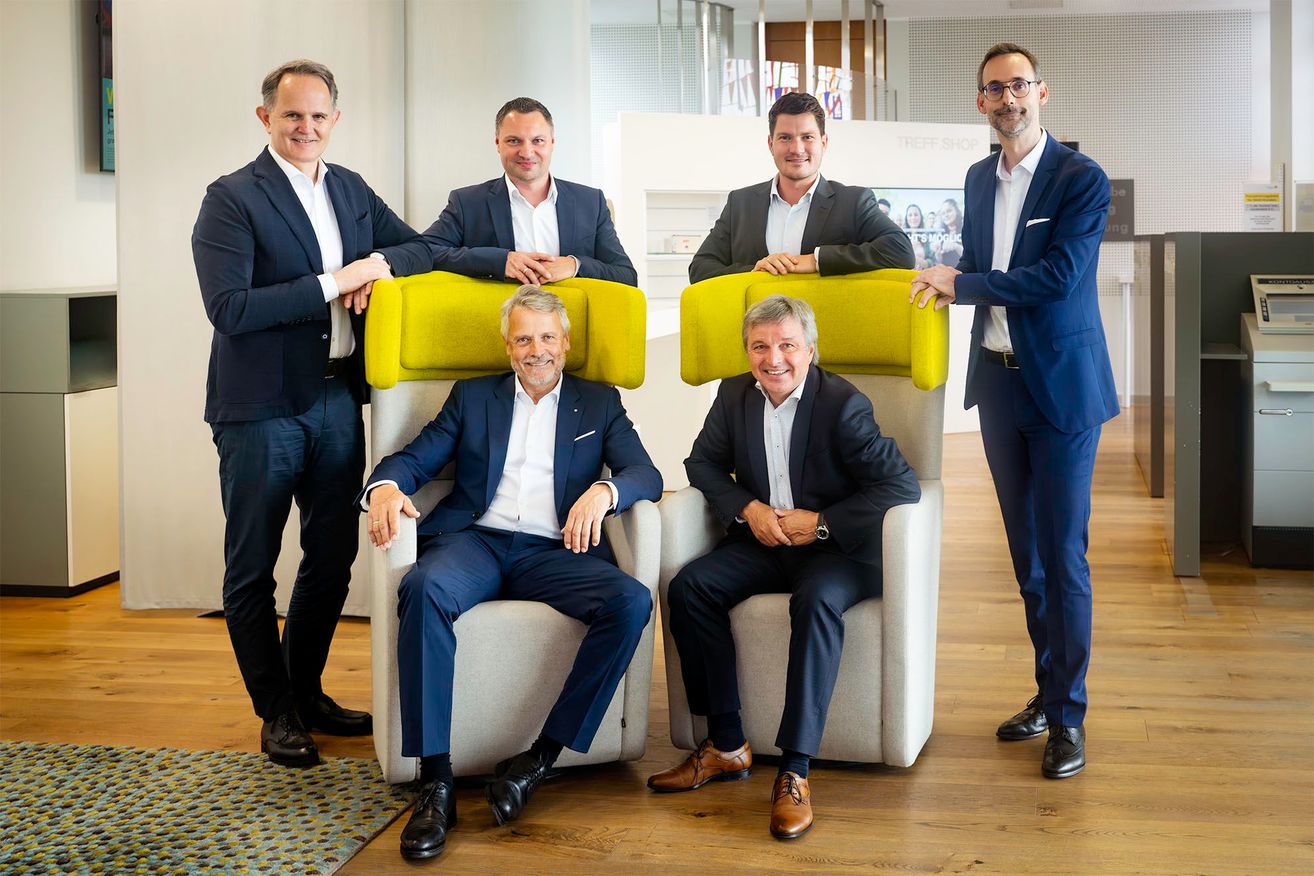 Five men in business suits pose for a photo in a modern office setting, with two sitting on yellow chairs and three standing behind them.