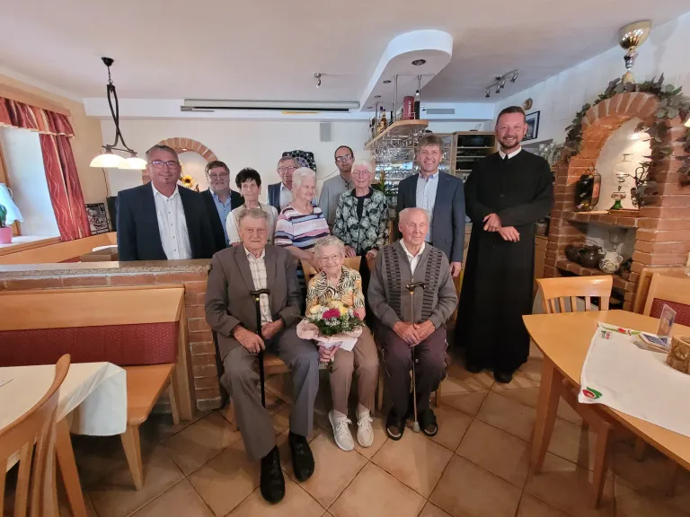 A group of elderly people and a priest are posing for a photo in a restaurant. The elderly people are sitting on chairs, while the priest is standing. The elderly woman in the middle is holding a bouquet of flowers. Behind them are a brick wall, a shelf, and a lamp.