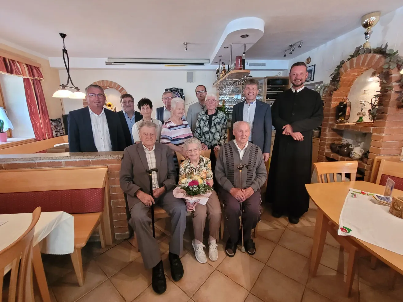 A group of elderly people and a priest are posing for a photo in a restaurant. The elderly people are sitting on chairs, while the priest is standing. The elderly woman in the middle is holding a bouquet of flowers. Behind them are a brick wall, a shelf, and a lamp.