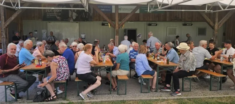 A group of elderly people are seated at tables in an outdoor restaurant, some are drinking and others are conversing. The tables are surrounded by wooden benches.