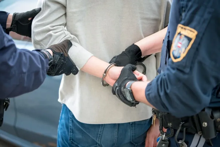 A person in handcuffs is being escorted by two police officers, one holding their left arm and the other holding their right.