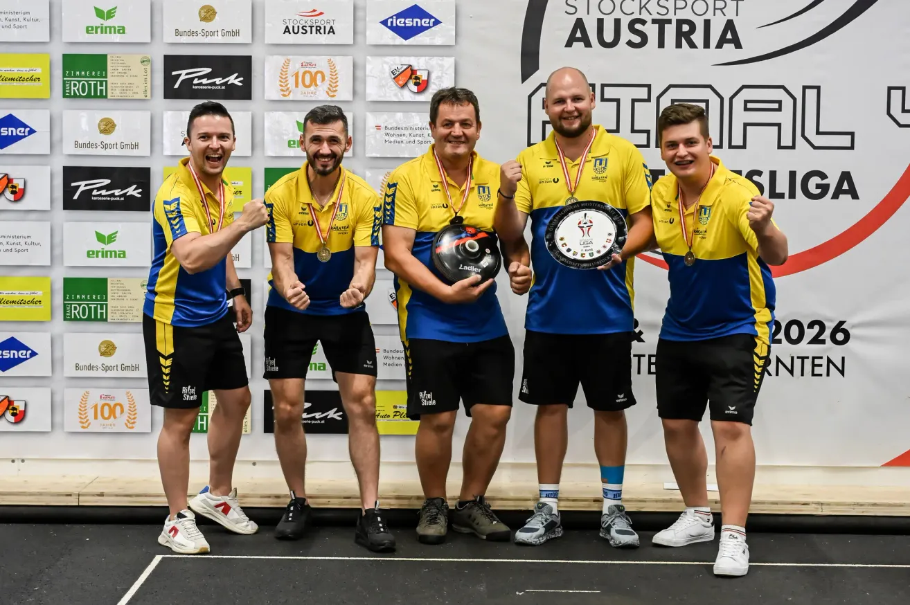 Five men in yellow and blue jerseys with medals around their necks are standing together and posing for a photo. They are holding a trophy and a helmet. Behind them is a wall with logos and text.