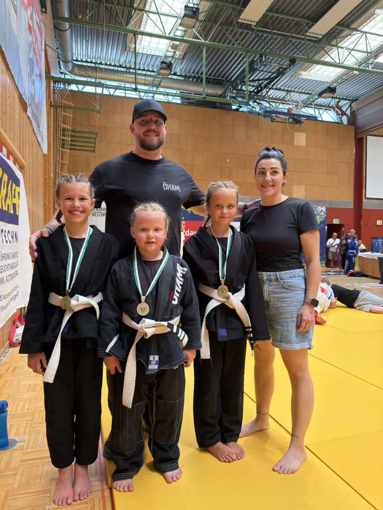 A group of young girls and a man in martial arts uniforms stand on a yellow mat, smiling for a photo. The man has his arms around two of the girls. They all wear medals around their necks. Behind them, a wooden wall with a banner and a projector screen is visible.