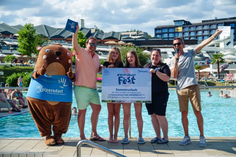 A group of people and a mascot pose for a photo in front of a pool, holding a banner for the #WeAreWater Fest.