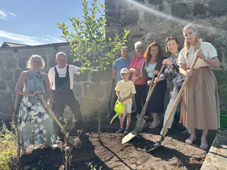 Eine Gruppe von Menschen pflanzt einen Baum in einem Garten. Sie halten Schaufeln und Gießkannen. Das Kind hält eine Gießkanne. Die Frau rechts lächelt. Die Frau links trägt ein Kleid. Der Mann in der Mitte trägt ein weißes Hemd. Die Mauer hinter ihnen ist aus Stein.