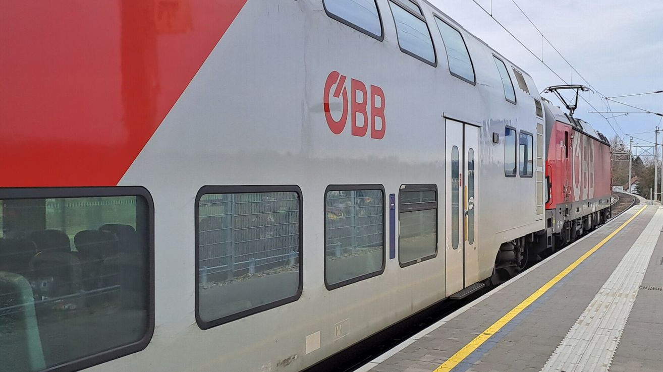 A silver train with red accents and the letters OBB on the side, parked at a station with a yellow line on the platform.